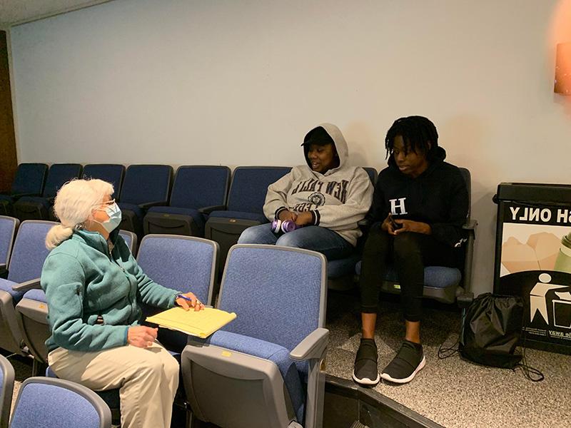 A woman with white hair and protective facemask turns around in her chair to talk with two female students sitting behind her. 