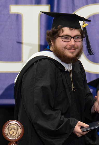 一个留着浓密胡须、戴着黑框眼镜、头戴黑色毕业帽、身穿黑色毕业礼服的男子微笑着与镜头外的人握手