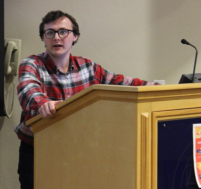 Male student with glasses and checked shirt making remarks while standing at a podium