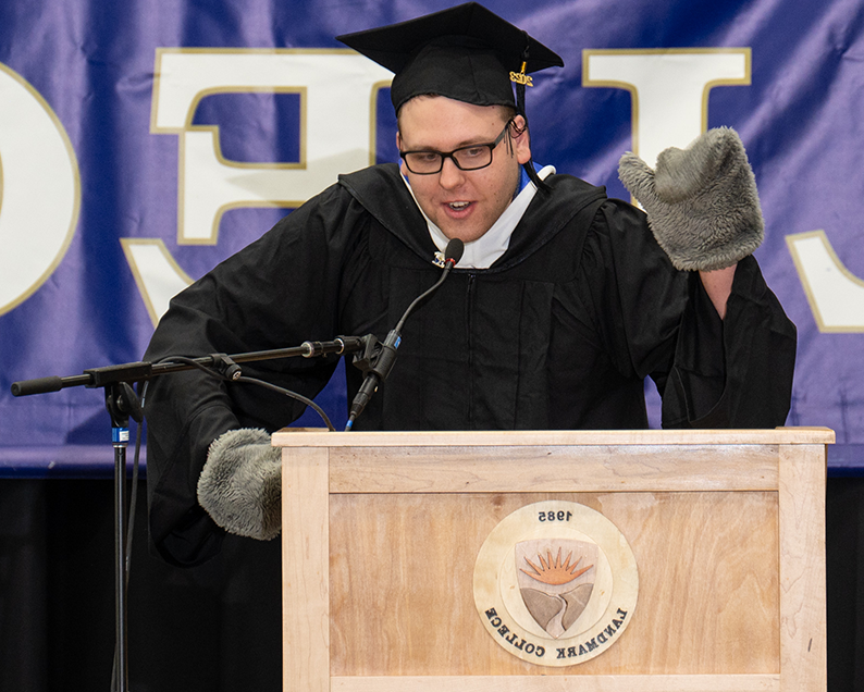一名戴着眼镜、戴着黑色毕业帽、身穿长袍的男学生站在讲台上，手里拿着一只灰色的脚蹼，向观众挥手致意