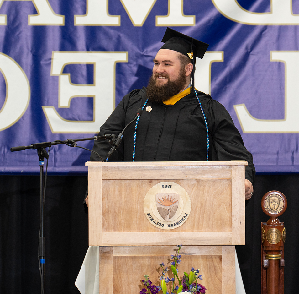蓄着棕色浓密胡须、头戴黑色毕业帽、身穿黑色毕业礼服的男子站在讲台上微笑